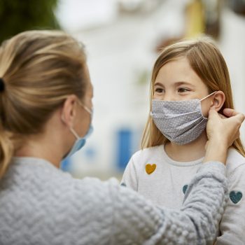A mother wearing surgical mask helps daughter putting home made face mask during coronavirus pandemic.